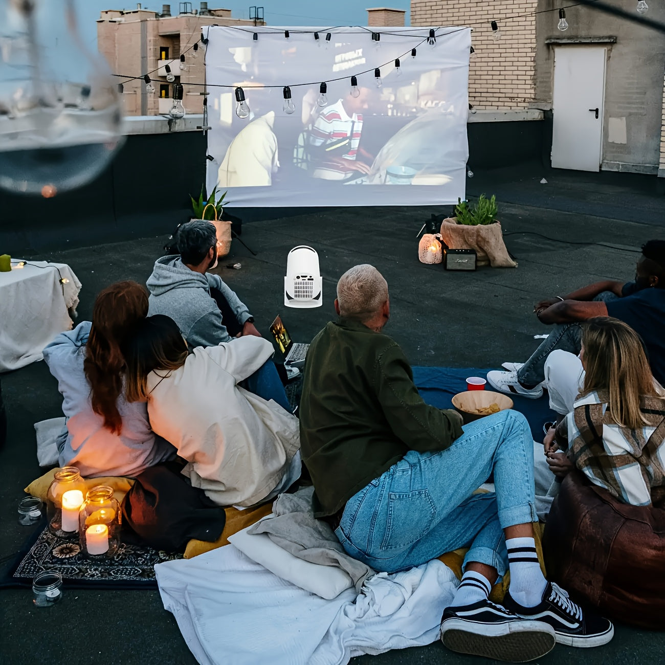 Mini projecteur Full HD 1080p projecting a movie onto a screen, with people sitting on blankets in an outdoor setting.